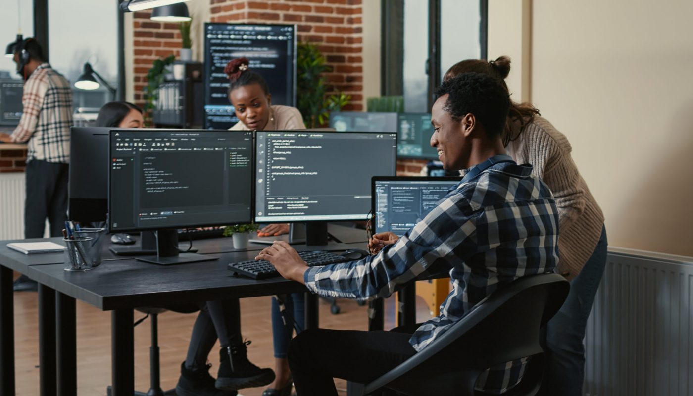 Software developer looking at running code on multiple screens takes off glasses and doing high five hand gesture with colleague programer. Programmers celebrating successful online cloud computing.